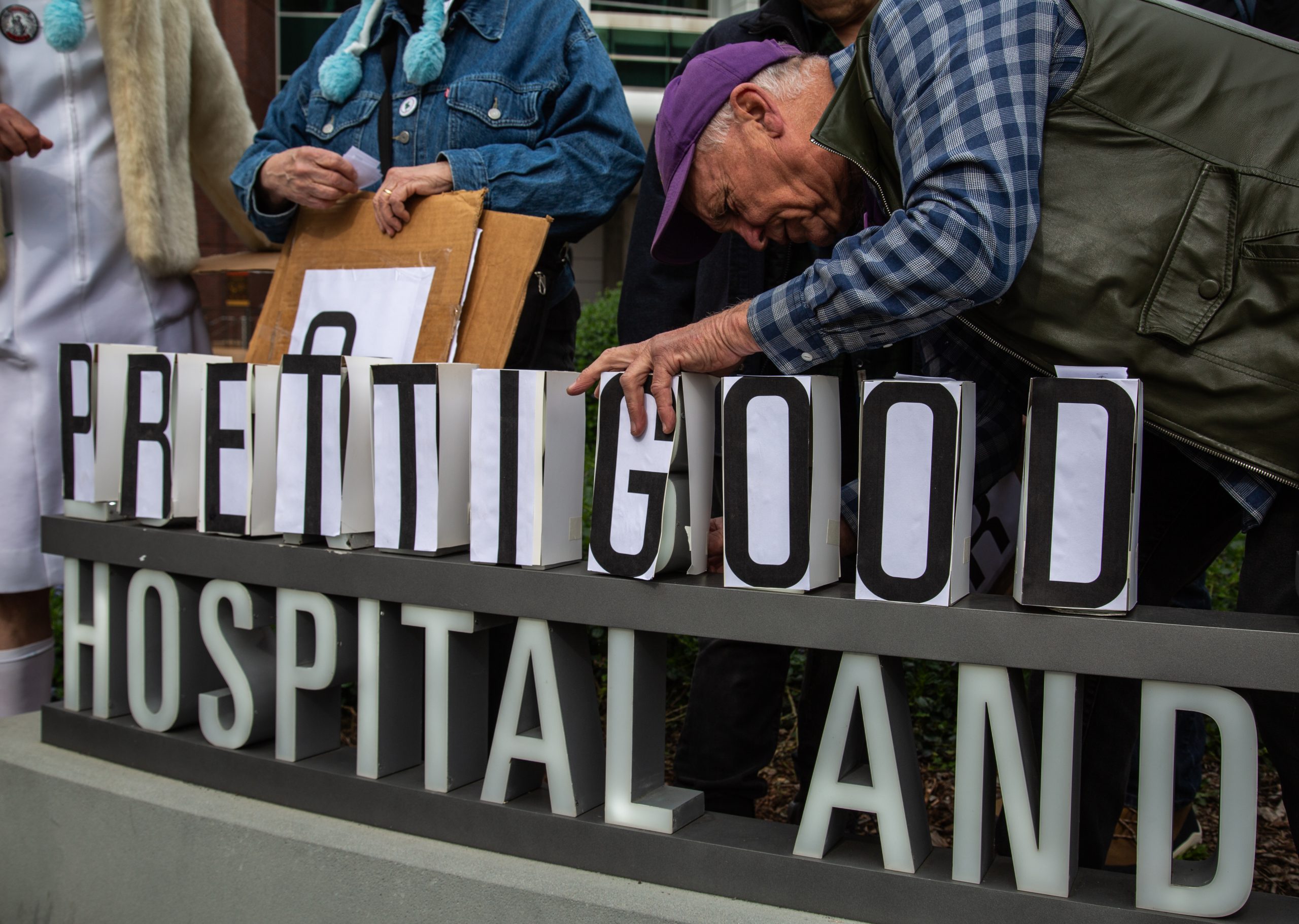 A group of people rearrange letters on a hospital sign to spell out "PRETTY GOOD" over the original sign.
