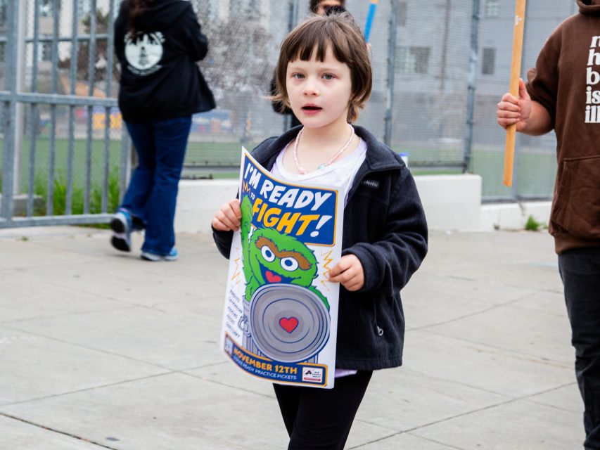 A young girl holds a sign that says "I'M READY TO FIGHT!" with an illustration of Oscar the Grouch, walking on a sidewalk near a fence.