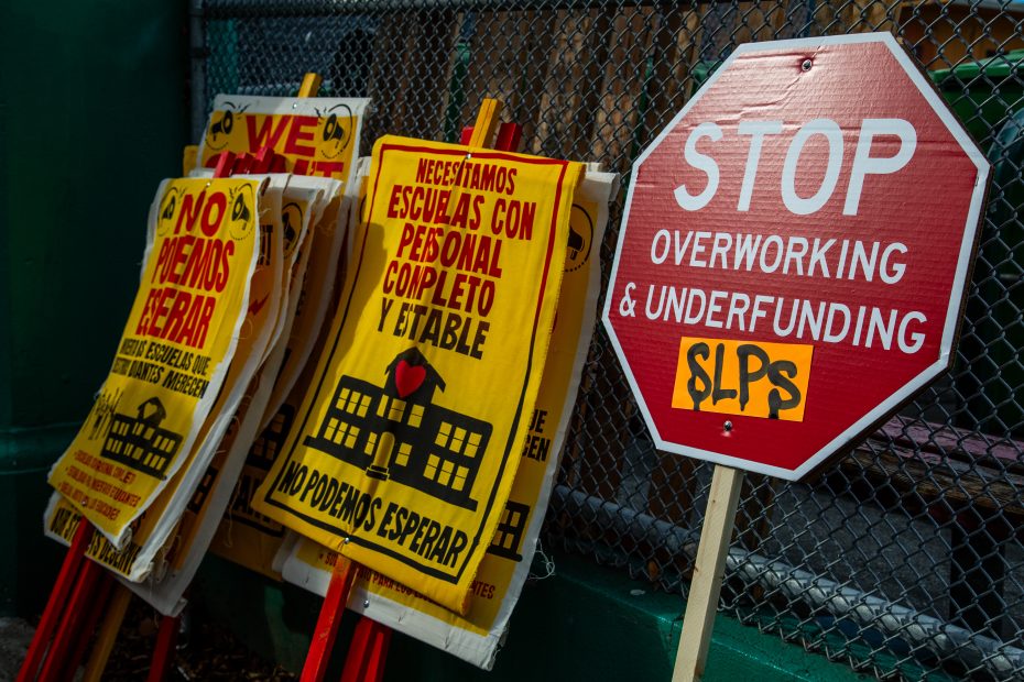 Protest signs in English and Spanish demand better staffing and funding for schools, with a stop sign reading "Stop Overworking & Underfunding SLPs" leaning against a fence.
