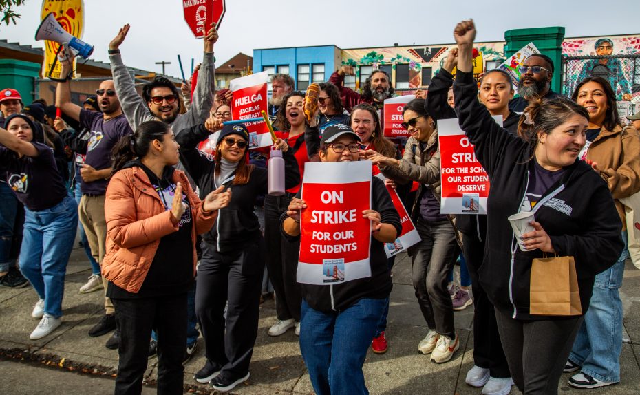 A group of people stand and cheer outside, holding signs that read “ON STRIKE FOR OUR STUDENTS,” participating in a strike or protest.