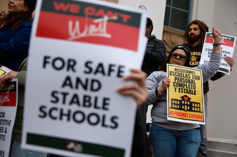 People stand at the steps of a school during a strike. The photo focuses on a person holding up their left hand in a fist while they hold a sign that reads "we can't wait" in Spanish with the other hand.