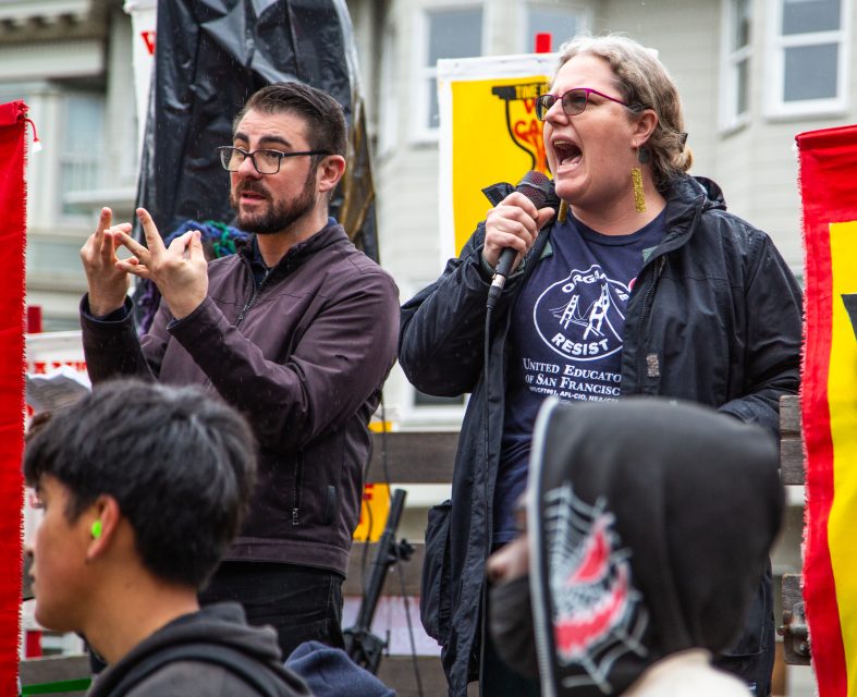 A woman speaks into a microphone at an outdoor event while a man beside her translates in sign language; people and colorful banners are visible in the background.