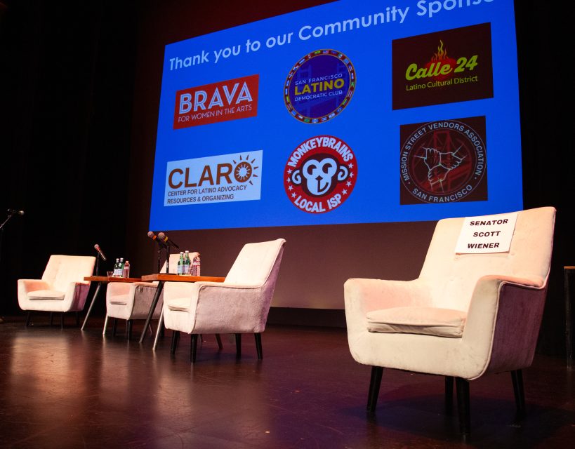 A stage with four empty chairs, a table with water bottles, and a reserved sign for Senator Scott Wiener; sponsor logos are displayed on a screen in the background.