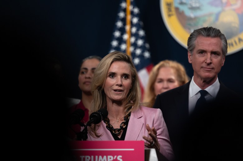 A person, with blonde hair and wearing a pink blazer, speaks in front of a podium while surrounded by other people. The California seal and the American flag can be seen in the distant background.