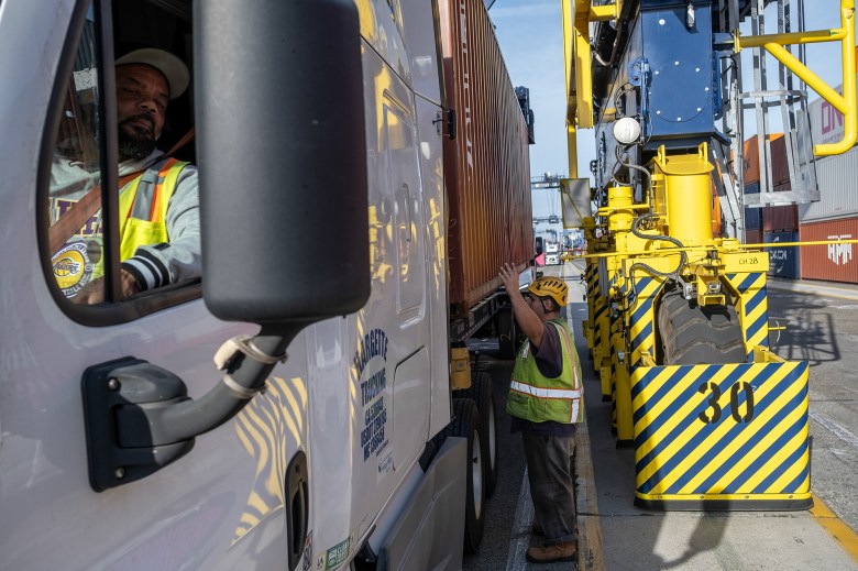 A person, wearing a yellow safety hat and vest and gray pants, stands next to a semi-truck while signaling with their left hand at a shipping terminal. The driver of the semi-truck uses their side mirror to see what the other person is doing.