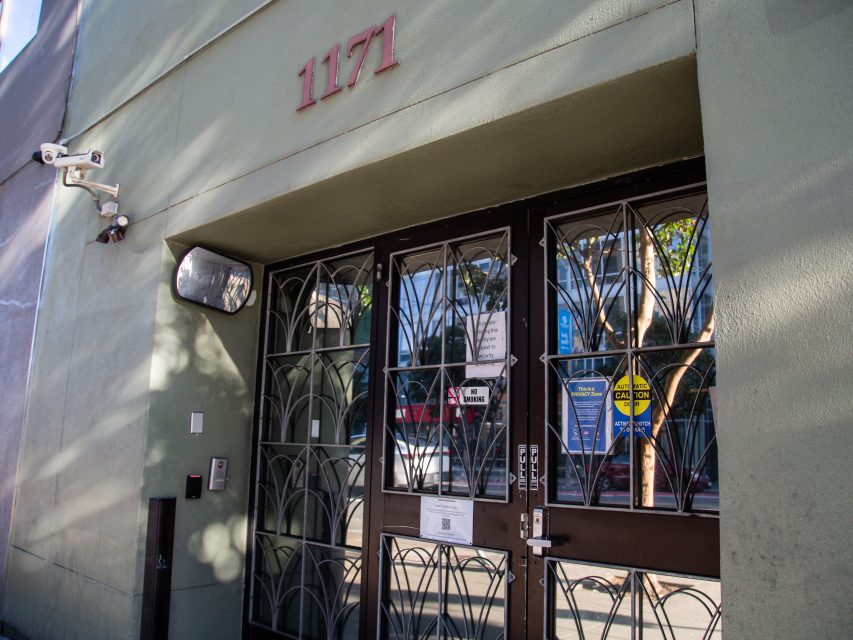 Entrance of a building with the address number 1171 above double doors, security cameras, a keypad, and various signs on the glass panels.