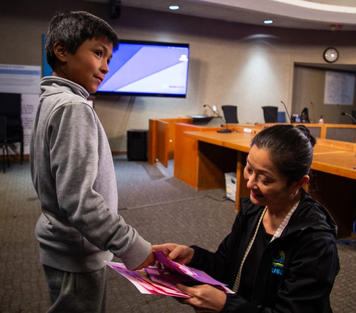 A woman kneels and hands colorful papers to a boy in a grey tracksuit inside a conference room with a large screen and wooden tables.