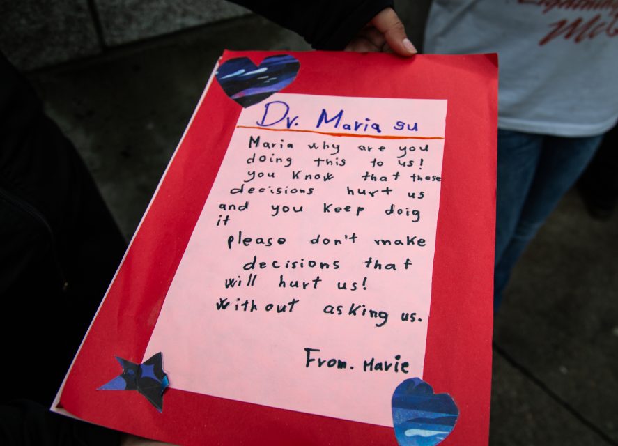 A hand holds a red card with a handwritten message addressed to Dr. Maria, asking her not to make hurtful decisions without consultation. Blue heart cutouts decorate the card.