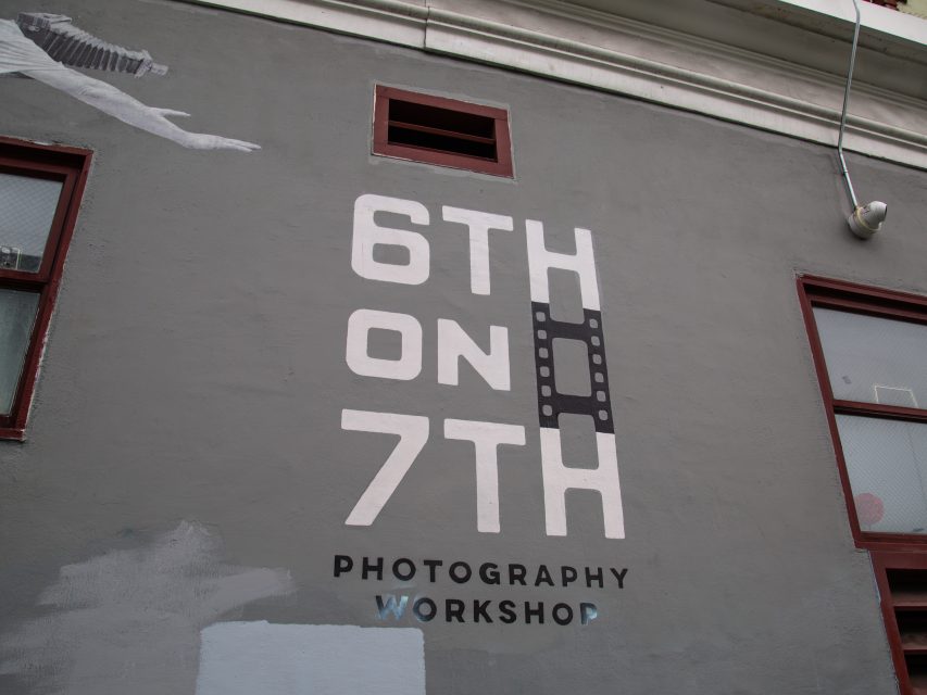 Gray wall with painted text reading "6th on 7th Photography Workshop," with the second "H" designed to resemble a filmstrip. Two windows and building details are also visible.