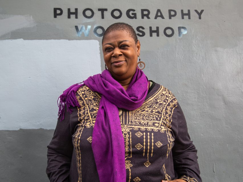A woman in a dark patterned top and purple scarf stands in front of a wall with the words "PHOTOGRAPHY WORKSHOP" painted on it.