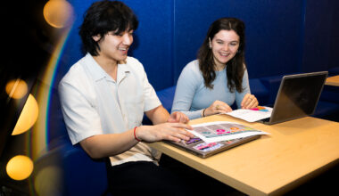 Dylan Kao and Lucy Paolini sit at a table reviewing colorful printed materials, with a laptop open, against a blue background with warm bokeh lighting on the left.