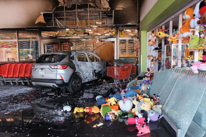 A burned SUV sits inside the entrance of a damaged store, its exterior charred and paint blistered, surrounded by soot-blackened walls and debris. Red shopping carts line the left side, while stuffed toys and shattered glass lie scattered across the wet floor in the foreground.