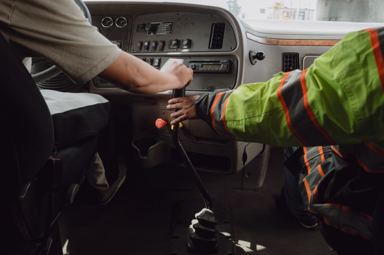 From behind a driver and passenger seat, the driver's hand is on the vehicle's gear stick while another arm in a yellow safety jacket extends out to guide the gear stick.