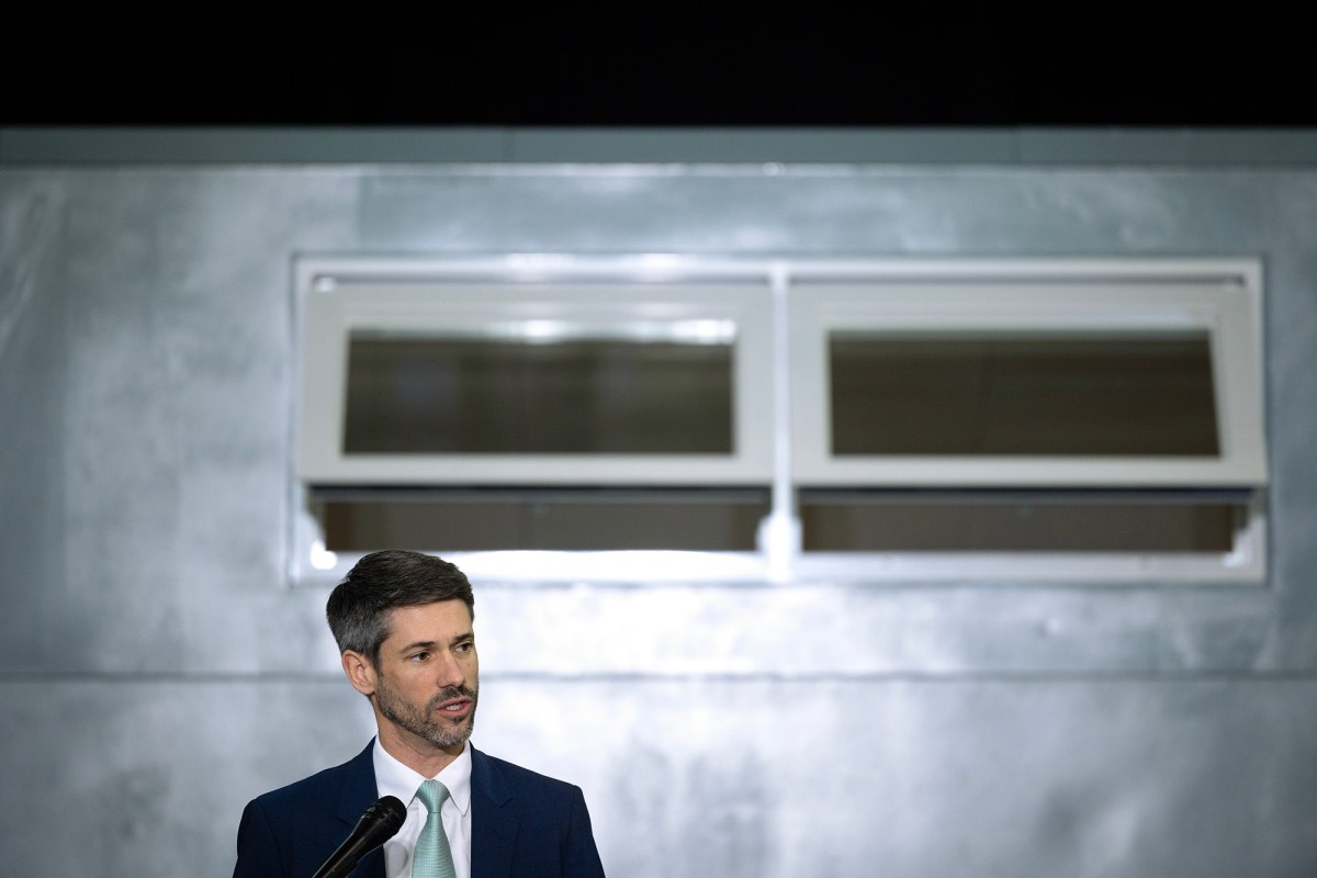 The image shows a person in a suit and tie, standing behind a microphone. They have have short, dark hair with some gray and a beard. The background features a metallic surface with a window above their head, which appears to be partially open. The person is speaking.