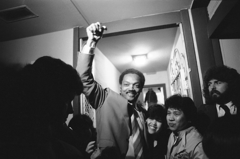 A black and white photo of a group of people gathering around Rev. Jesse Jackson as he smiles and raises a fist up in the air.
