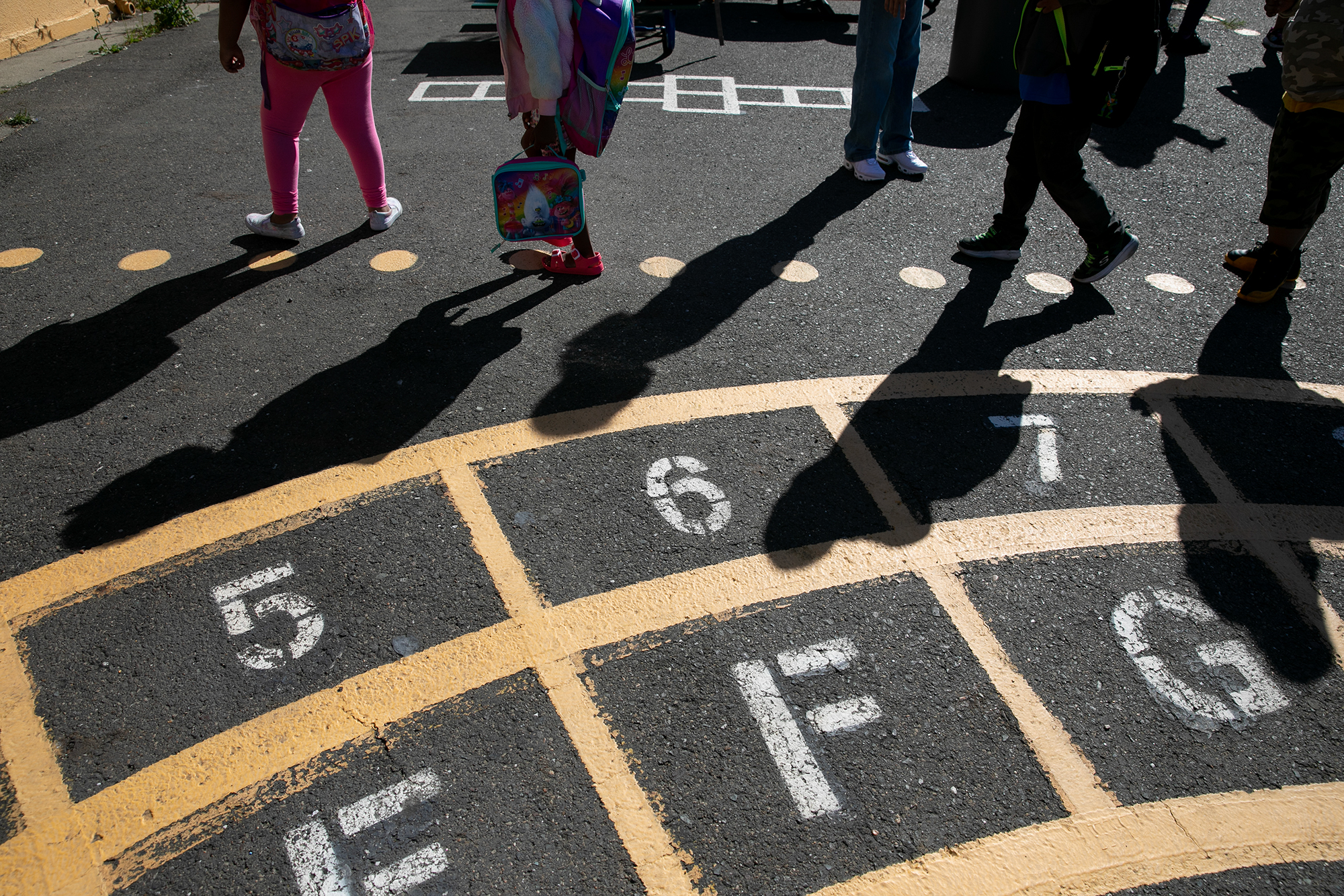 The bottom half of several children on a concrete playground with yellow chalk outlining numbers and letters is shown.