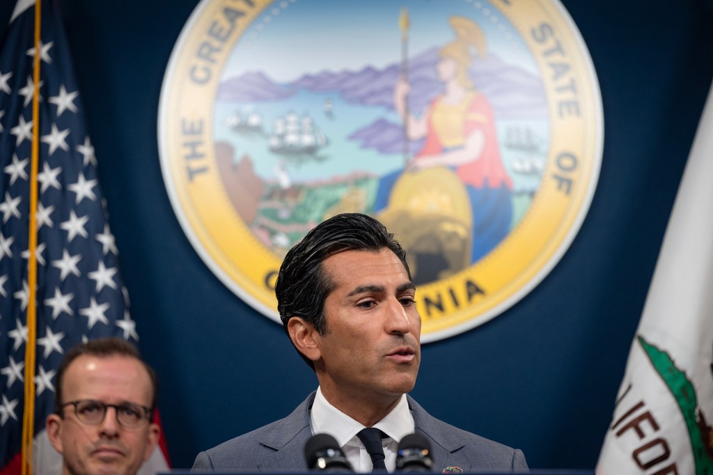 A suited speaker stands at a podium in front of the California state seal, with U.S. and California flags on either side, speaking into microphones while another person listens behind them.