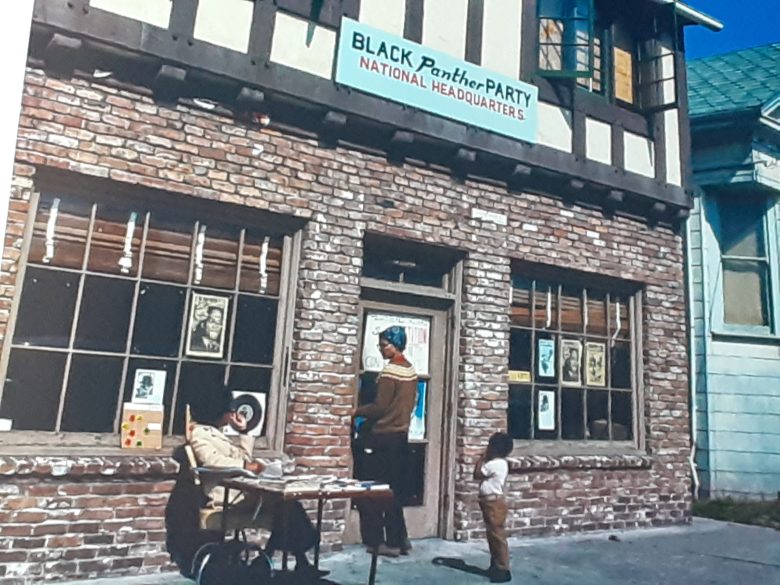 A sign reading "Black Panther Party National Headquarters" hangs above a door at a home, its windows filled with posters, as three people stand outside