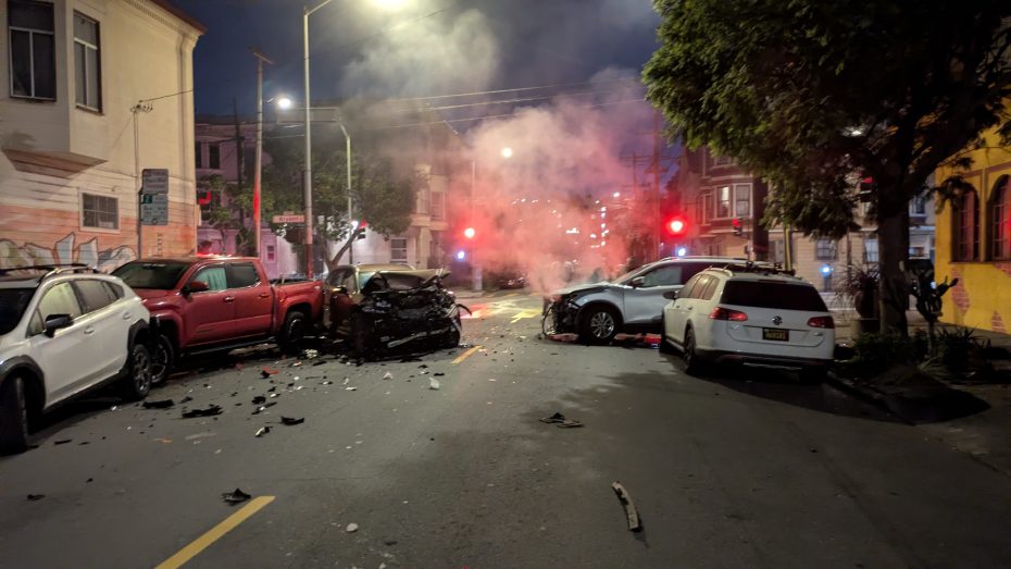 Several cars are damaged in a night-time street collision, with debris scattered on the road and smoke rising from one vehicle.