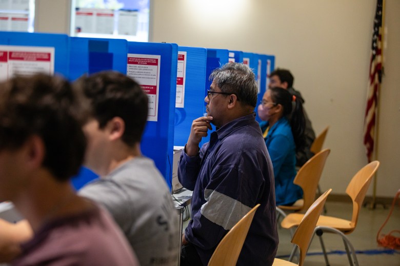 A side view of a row of voters sitting in front of voting machines separated by blue dividers. The photo focus on a voter who rest their right hand on their chin as they contemplate their voting selections.