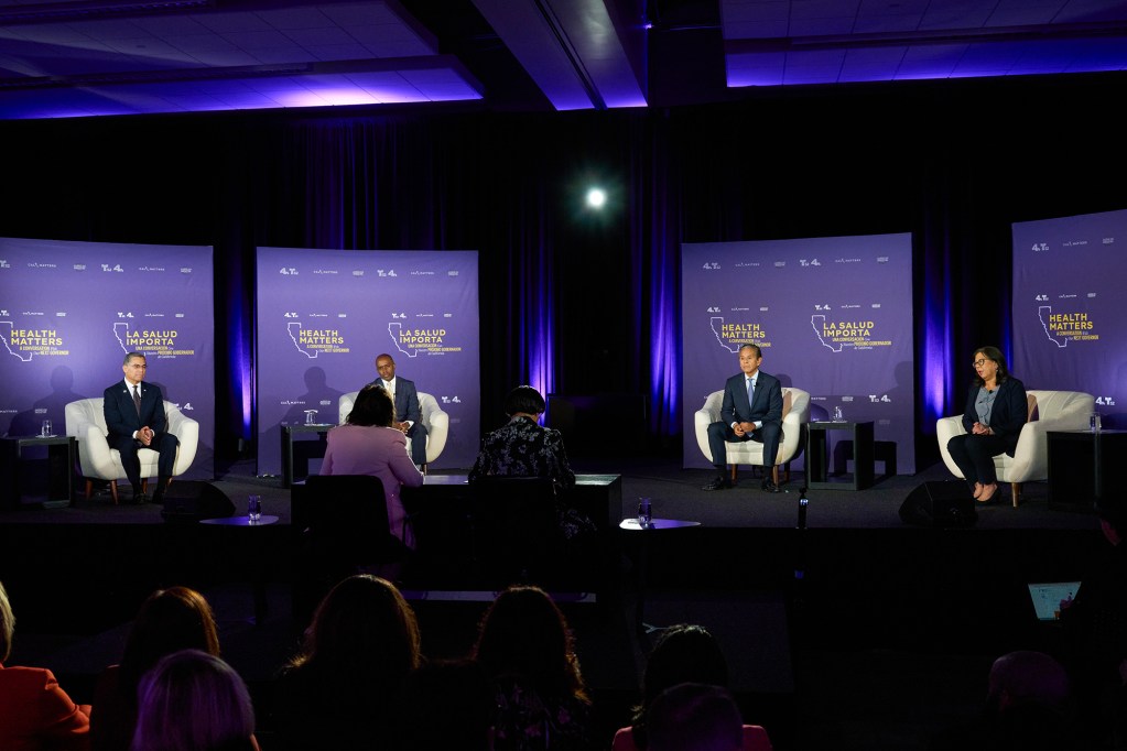 Four speakers sit onstage in armchairs for a moderated panel discussion, with an audience watching from the foreground. Behind them are purple backdrops reading “Health Matters” and “La Salud Importa,” lit by spotlights.