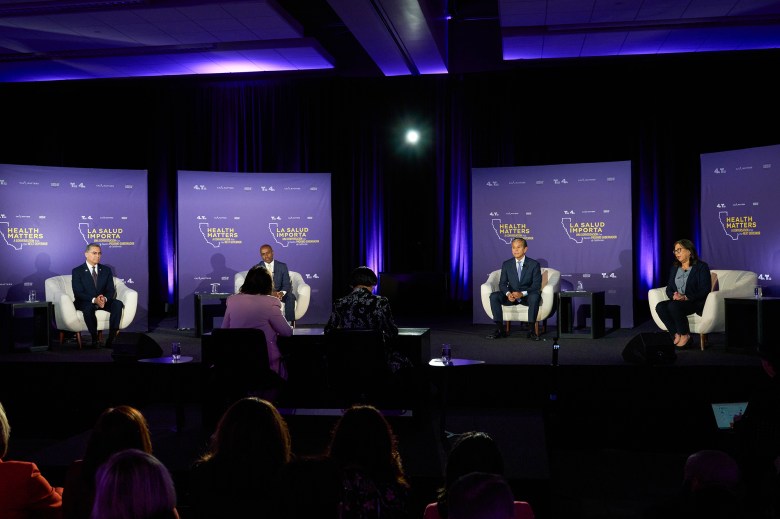 Four speakers sit onstage in armchairs for a moderated panel discussion, with an audience watching from the foreground. Behind them are purple backdrops reading “Health Matters” and “La Salud Importa,” lit by spotlights.
