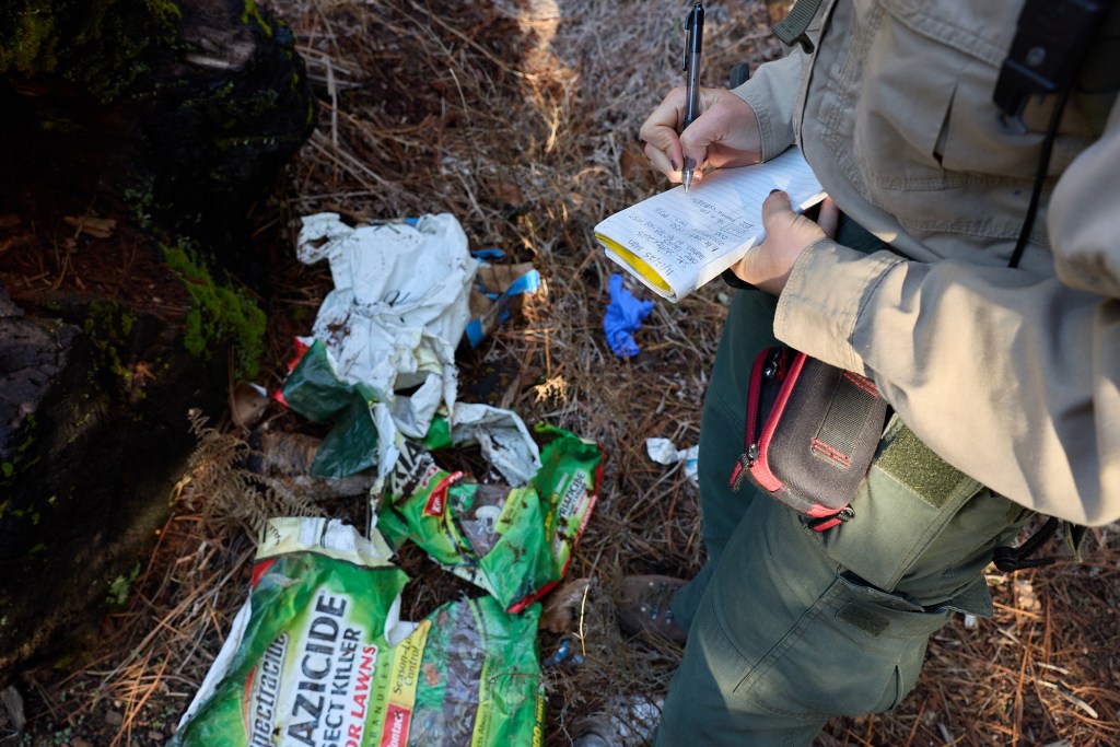 A person in brown hiking gear uses a pen to write in a small notebook at their waist as they look down at empty fertilizer bags in a grassy area of a forest.