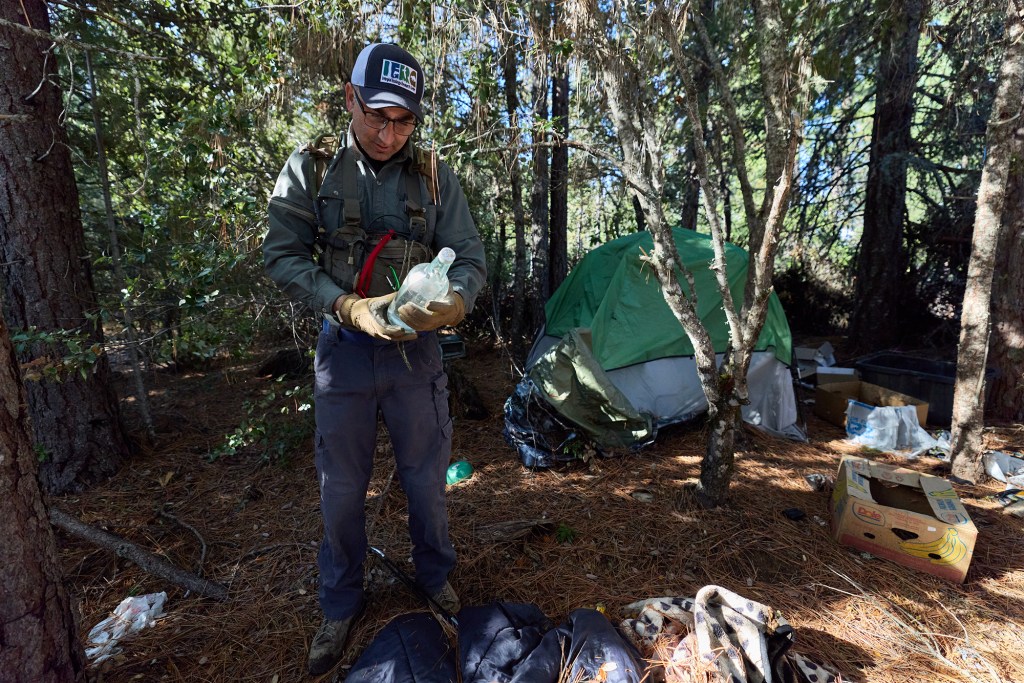 A person wearing hiking gear and a baseball cap stands in the center of a wooded area with a makeshift camp, holding a glass bottle. Behind them, a tent stands on a grassy area, with trash scattered across the campsite on the forest floor.