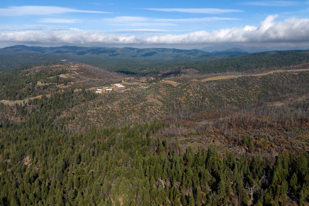 An aerial view of Post Mountain in a very densely forested area with pine trees along a mountainside and buildings in the background.