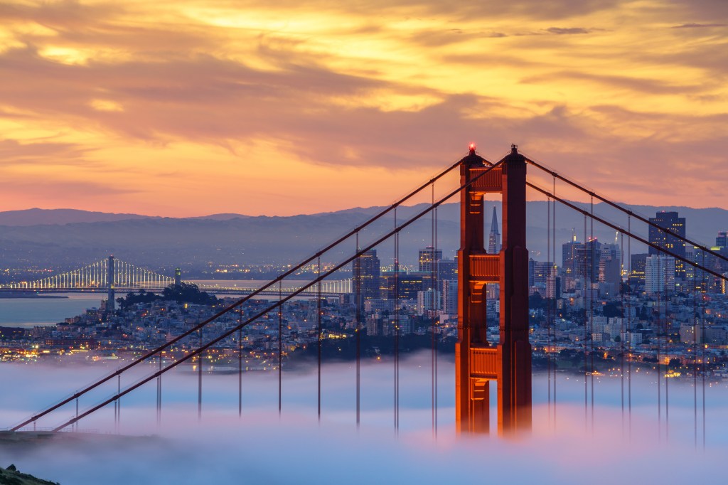 The Golden Gate Bridge at sunrise with low fog obscuring the bottom, revealing the San Francisco cityscape and the Bay Bridge.