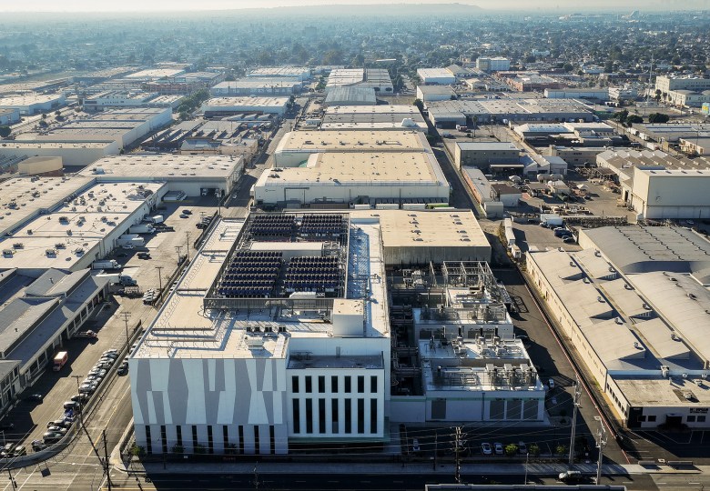 An aerial view shows a large data center complex surrounded by warehouses and industrial buildings in a dense urban area. The main structure has a white facade with vertical window slits and a flat roof filled with cooling units, pipes, and mechanical equipment, while streets, parked cars, and power lines run along the perimeter. A hazy cityscape stretches into the distance beyond the industrial zone.