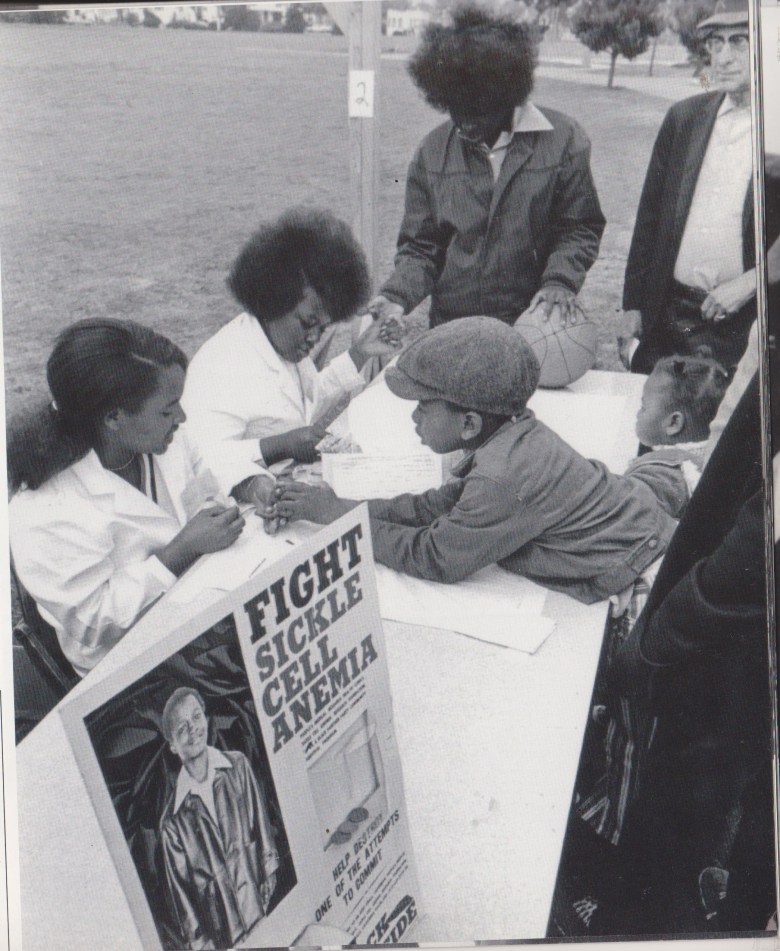 A black and white photograph depicts people sitting at a table, with a poster that reads "fight sickle cell anemia" in the foreground