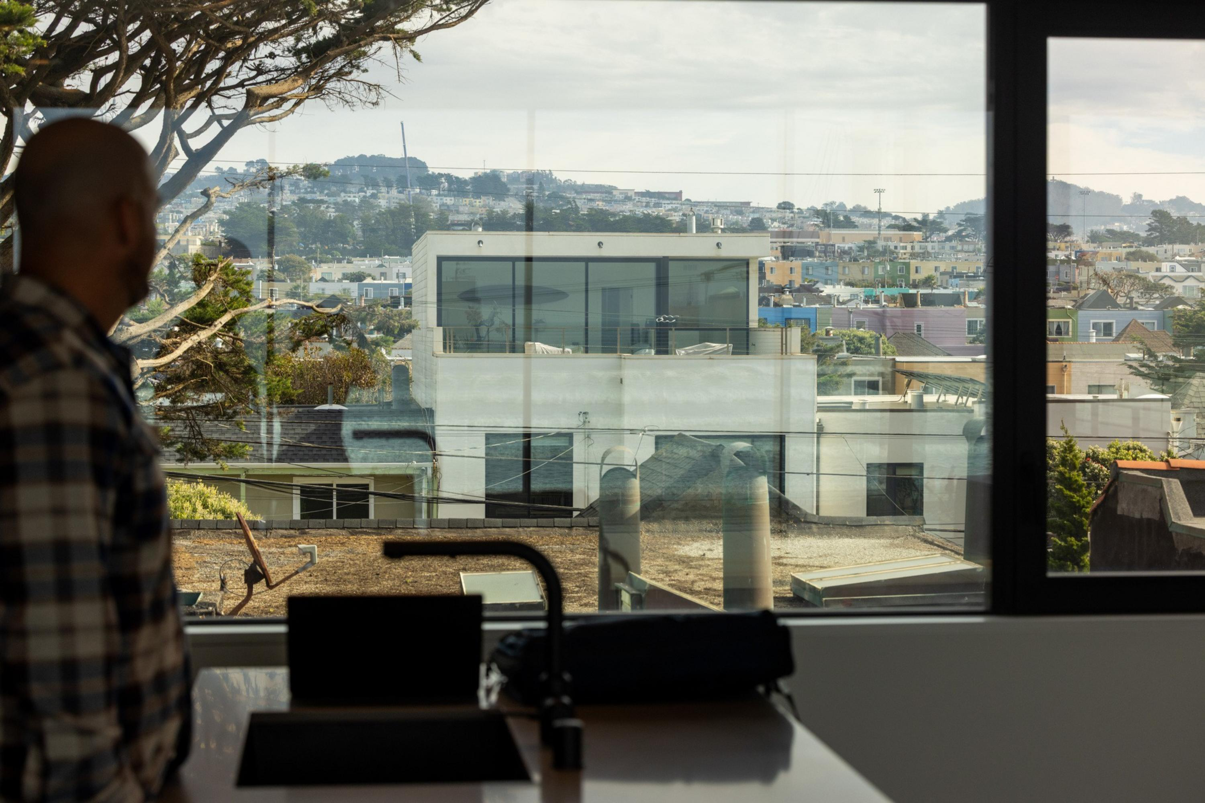 A man with a bald head and plaid shirt looks out a large window at a cityscape with modern buildings and hills in the distance.