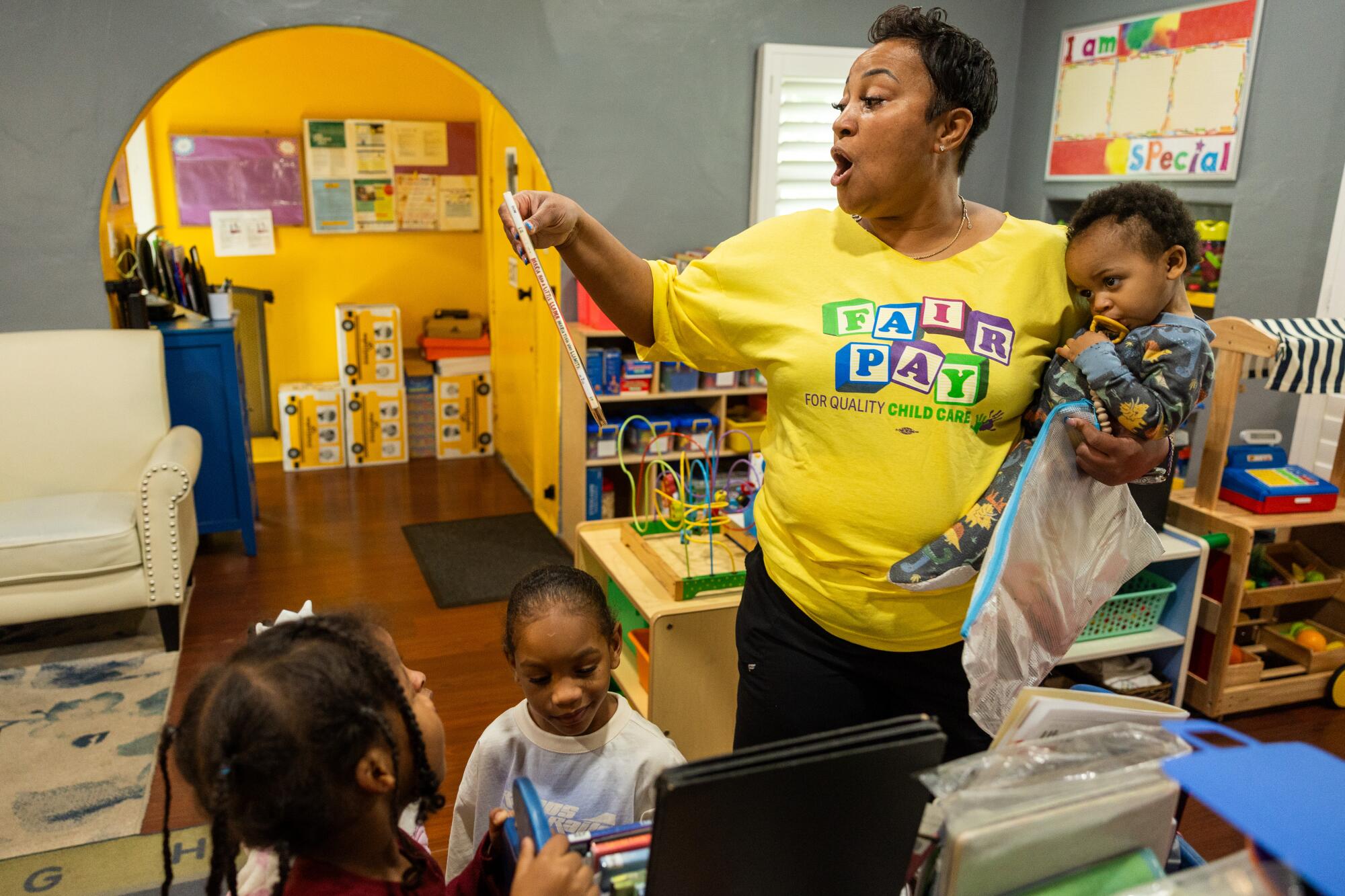 A child-care provider holds a child as she picks out a book to read from a shelf at her family childcare home.