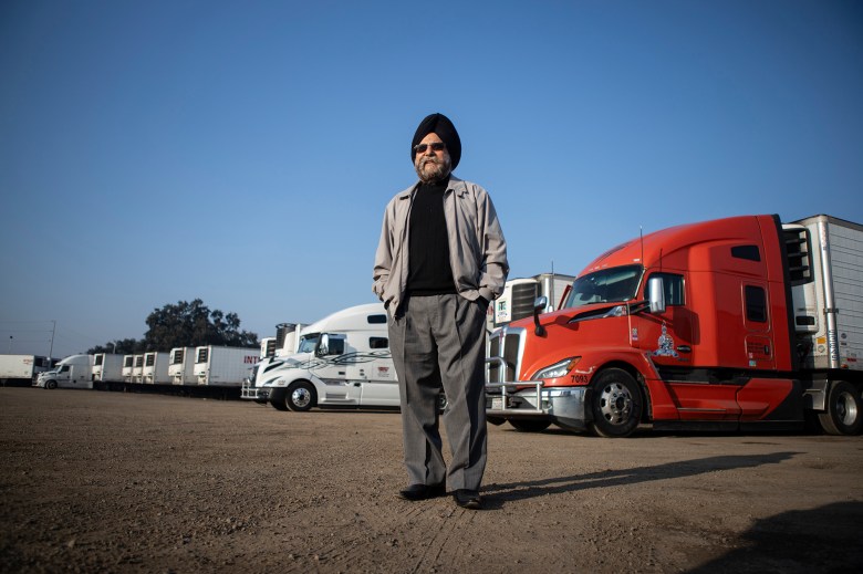 A person wearing a black turban, sunglasses, and a light jacket stands with hands in their pockets in a dirt lot lined with semi-trucks, including a bright red truck in the foreground and several white trucks stretching into the background under a clear blue sky.