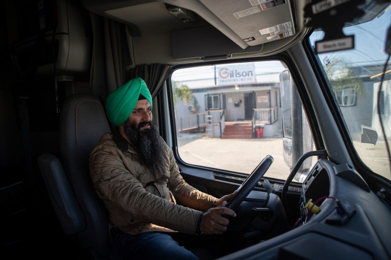 A truck driver wearing a green turban and quilted jacket sits behind the steering wheel inside a semi-truck cab, looking ahead as a trucking company office building is visible through the windshield.