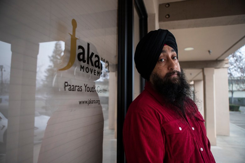 A person wearing a black turban and red jacket stands beside a glass door displaying the Jakara Movement and Paaras Youth Center logo, looking toward the camera along a covered walkway outside the building.