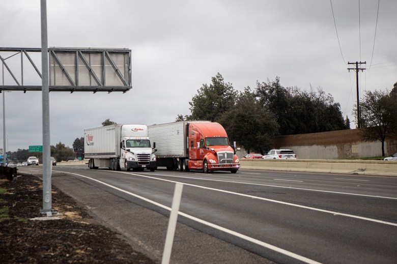 Two semi trucks, one white and one red, drive side by side along a multi-lane highway beneath an overcast sky, with trees, power lines and passing cars in the background.