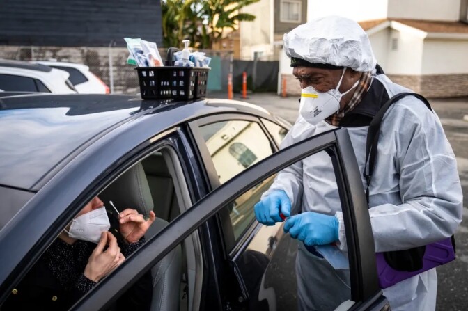 A person in a smock and a facemask administers a COVID-19 test to a woman in a black car. 
