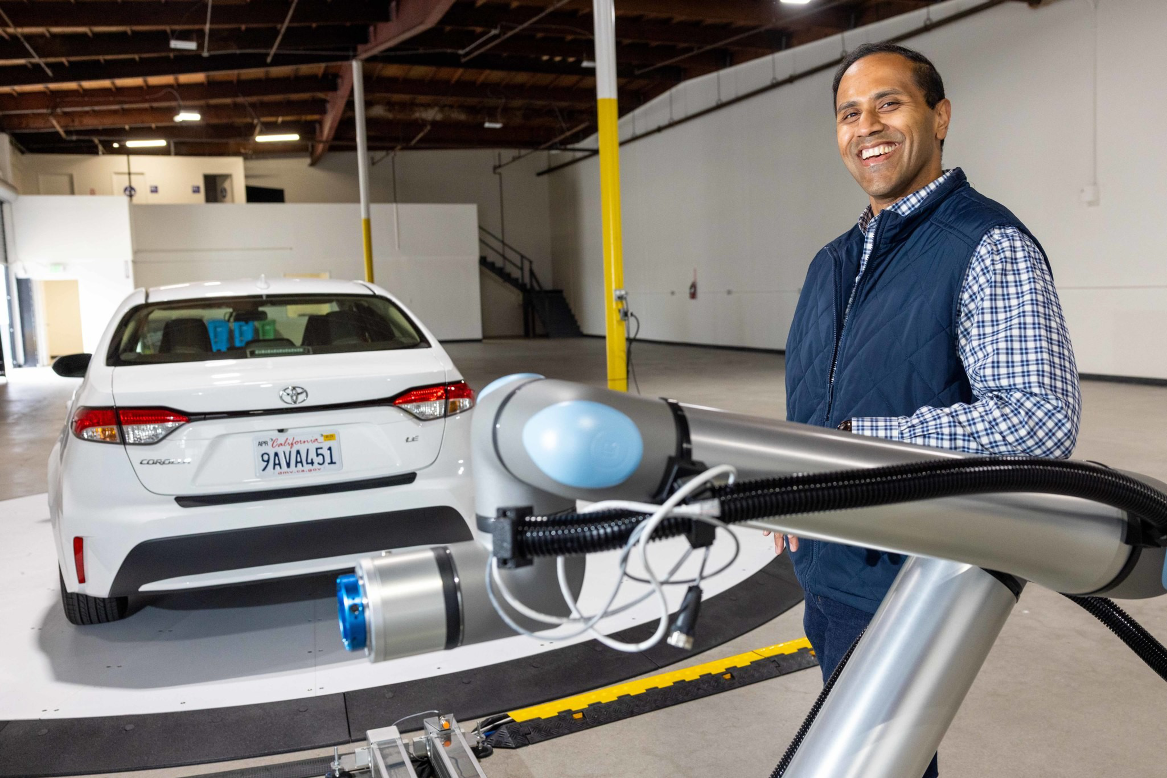 A man in a blue vest smiles beside a robotic arm, with a white Toyota car in a spacious indoor facility.