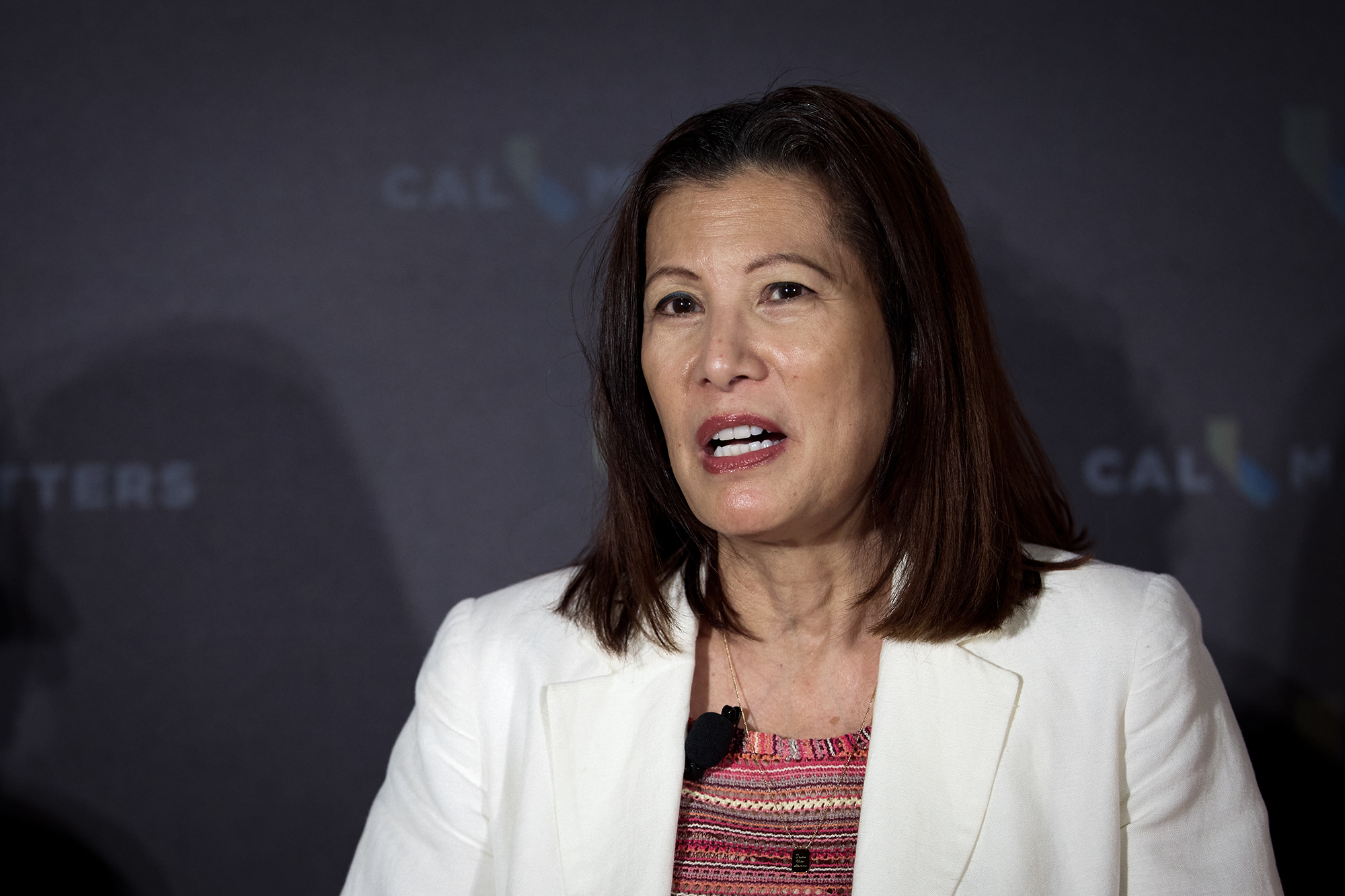 A person in a white blazer and patterned top speaks during an onstage interview, wearing a small lapel microphone, with a dark, softly lit backdrop behind them showing faint CalMatters logos.