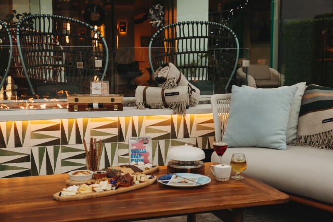A cosy set up, with a sofa, rattan chairs and a fire, with a table holding a cheese and charcuterie board and various drinks