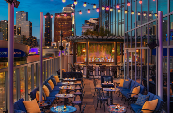 A rooftop bar at night, with lit up buildings in the background and fairy lights all around; in the middle are comfy seats and tables