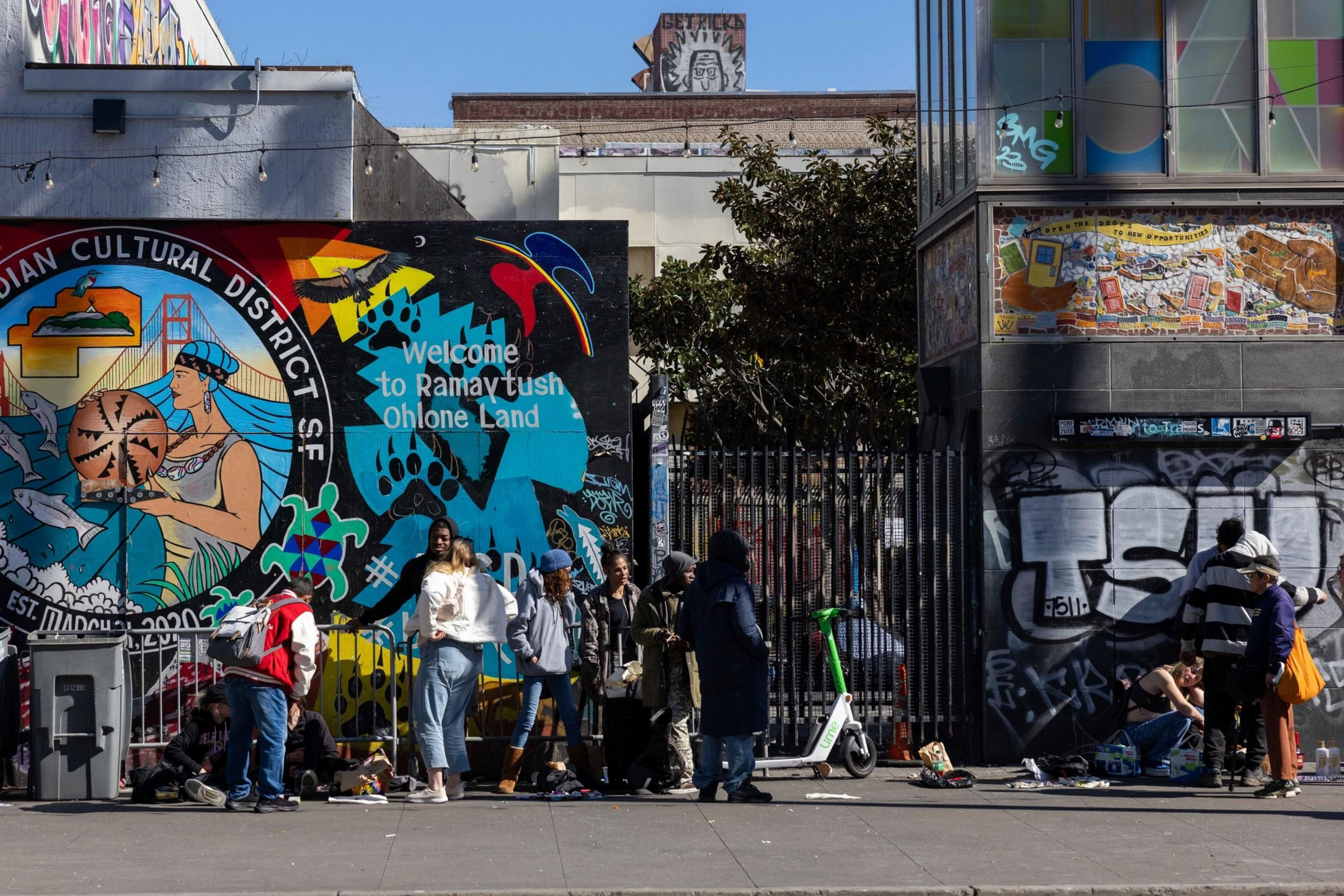 A mural on a city wall displays vibrant Indigenous imagery, with people gathered below it. Graffiti covers nearby surfaces, and one person stands by a scooter.