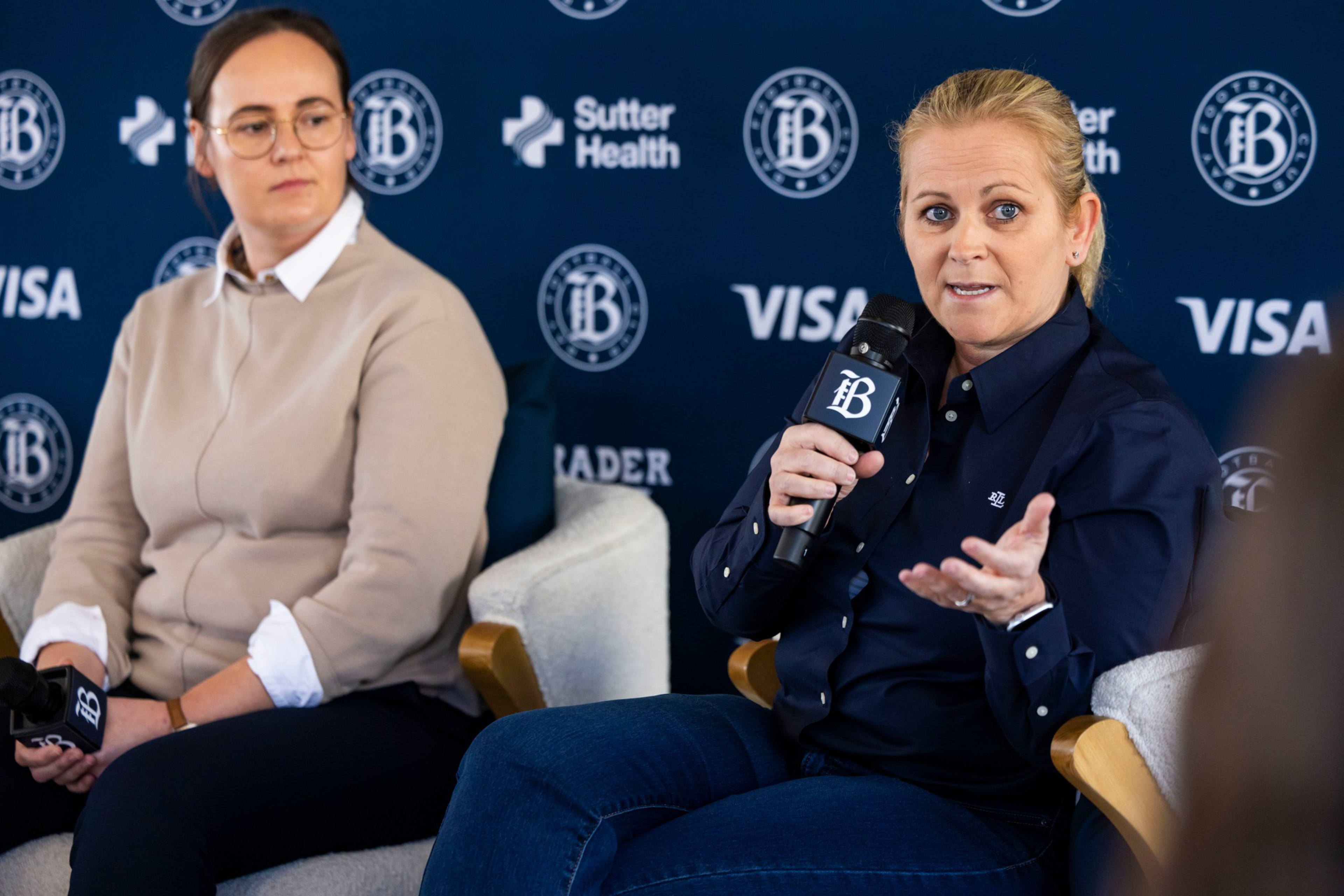 Two women sit in armchairs against a backdrop with logos, one speaks into a microphone while gesturing, the other listens holding a microphone in her lap.