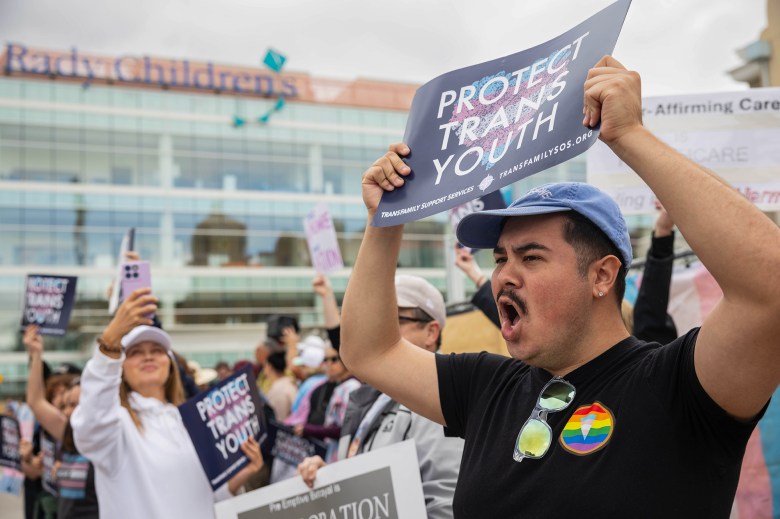 A person holds up a blue sign that reads "protect trans youth" as they stand next to other protesters in front of a hospital building.