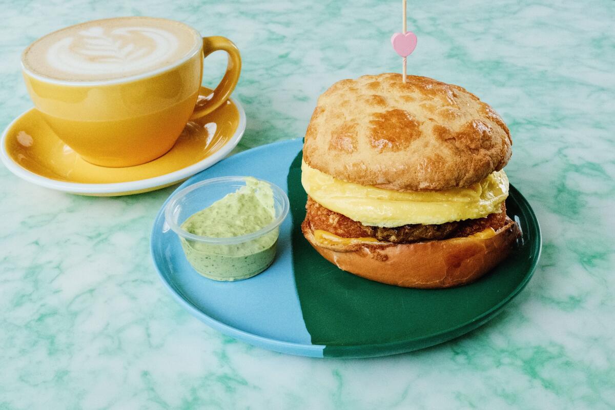 A pineapple-bun breakfast sandwich on a plate next to a latte in a yellow mug on a patterned green table