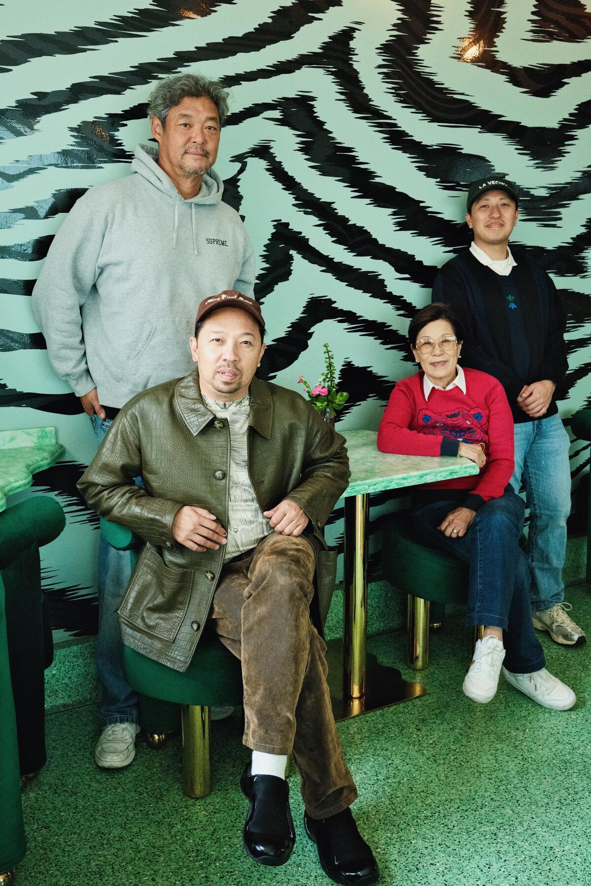 Two men stand while a man and a woman sit in a dining room against a black-and-green patterned wall