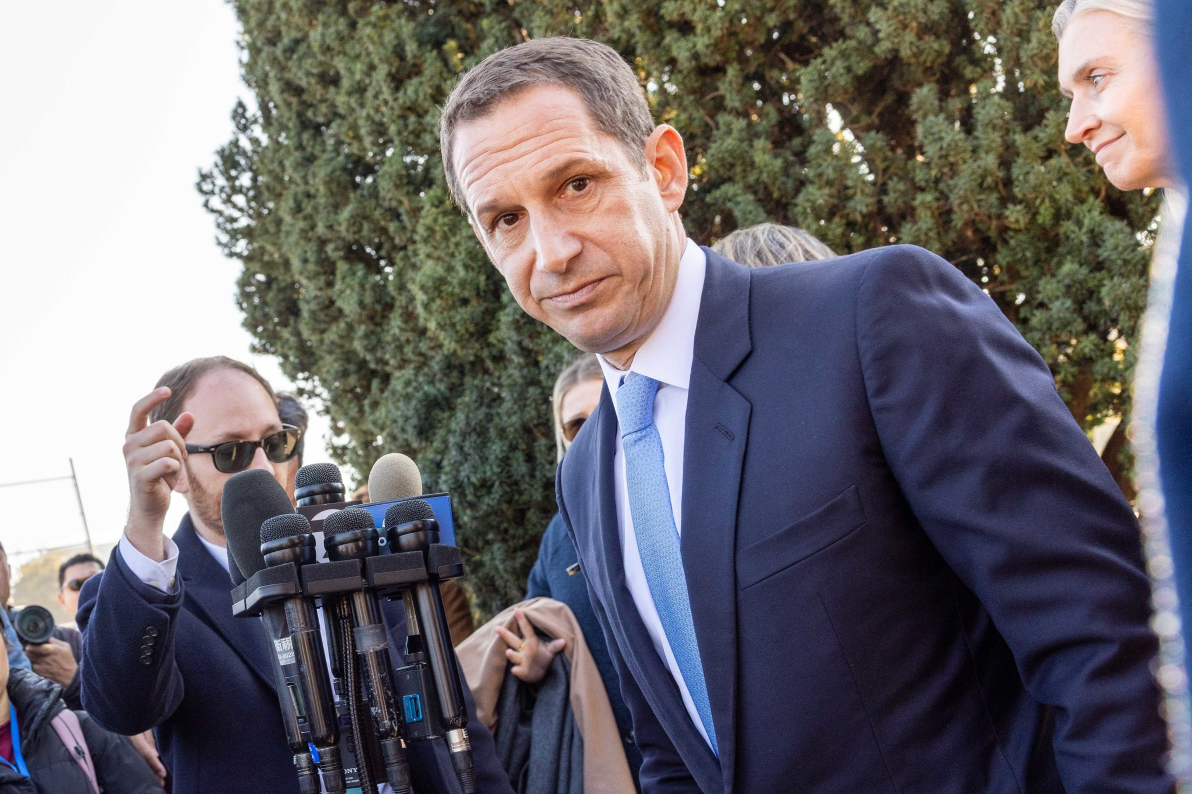A man in a blue suit and light blue tie leans toward several microphones, while another man wearing sunglasses and a group of people stand behind him.