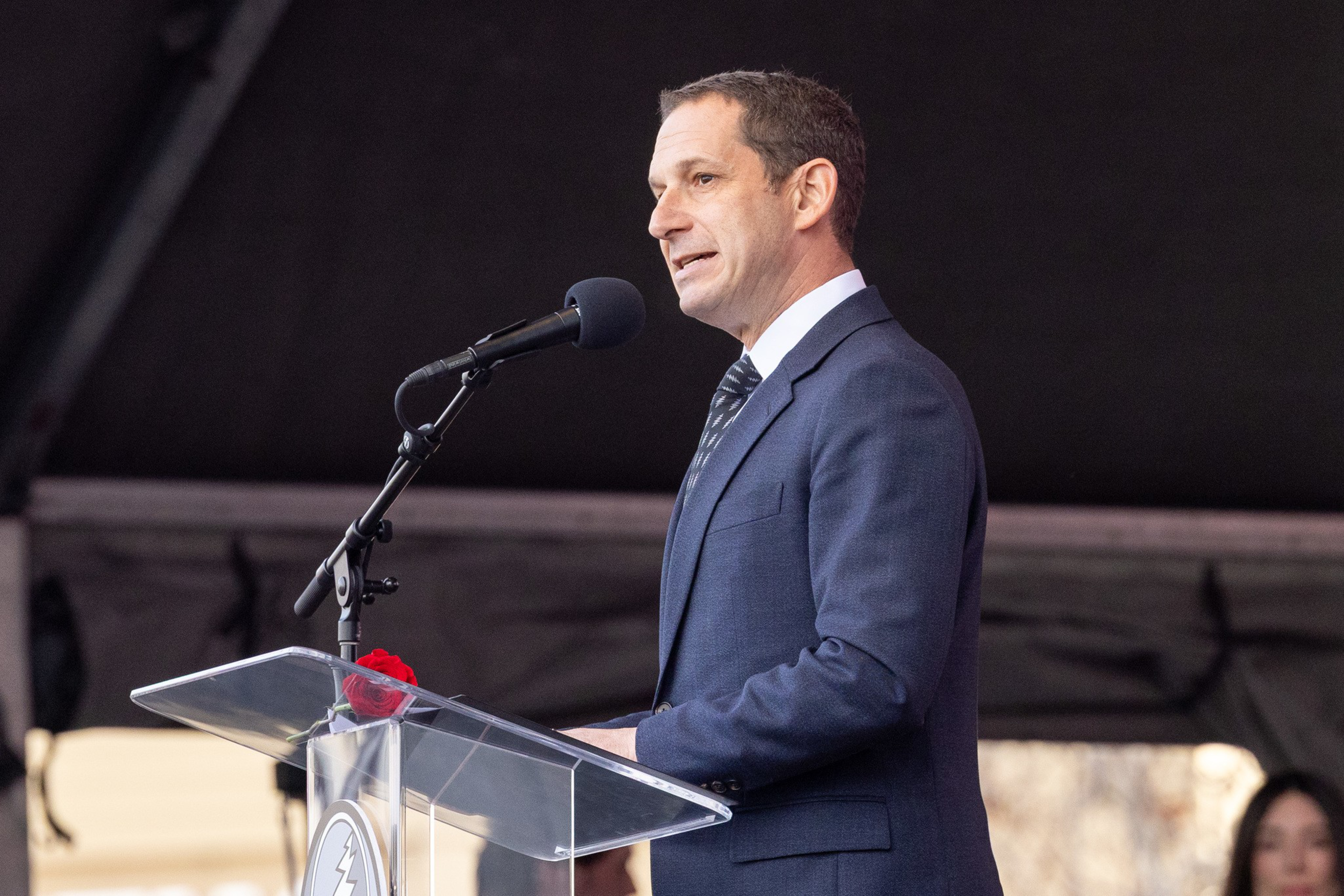 A man in a dark suit and striped tie stands behind a clear podium speaking into a microphone with a small red flower attached.